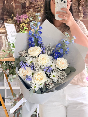 A person holding a large bouquet of flowers, including cream roses, blue delphiniums, white daisies, and lavender, wrapped in light grey paper. The person is taking a mirror selfie in a shop setting.