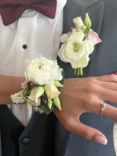 A close-up of a formal corsage and buttonhole set featuring a white rose wrist corsage and matching lapel boutonniere on a man in a grey suit jacket and white shirt.
