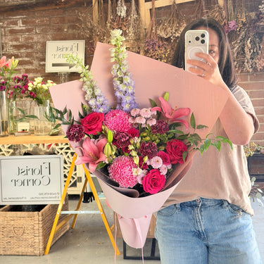 A person taking a mirror selfie in a florist shop, holding a large bouquet of pink and red flowers wrapped in light pink paper. The shop has a rustic brick wall background with dried floral arrangements.