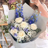 A person holding a large bouquet of flowers, including cream roses, blue delphiniums, white daisies, and lavender, wrapped in light grey paper. The person is taking a mirror selfie in a shop setting.