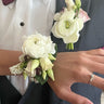 A close-up of a formal corsage and buttonhole set featuring a white rose wrist corsage and matching lapel boutonniere on a man in a grey suit jacket and white shirt.